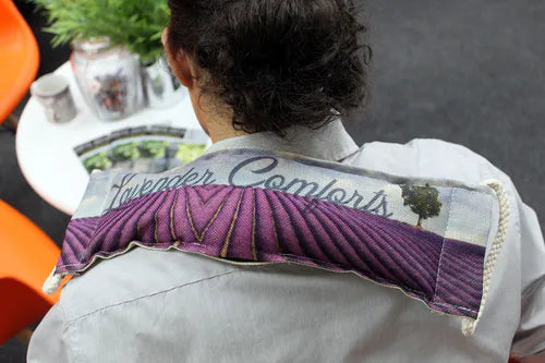 A person wearing a striped shirt with a lavender-themed wheat bag draped across their shoulders. The bag features a purple lavender field design with the words “Lavender Comforts,” providing a soothing and aromatic relaxation aid.
