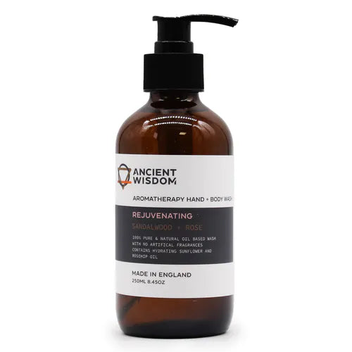 Brown plastic pump bottle of Ancient Wisdom Aromatherapy Hand + Body Wash. The white and black label highlights the "Rejuvenating" variant with sandalwood and rose. The wash is described as 100% pure and natural oil-based, free from artificial fragrances, enriched with hydrating sunflower oil and rosehip oil. The bottle contains 250ml (8.45oz) and is made in England.