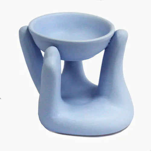Light blue ceramic oil burner shaped like a sculpted hands supporting a round shallow bowl for holding essential oils or wax melts.