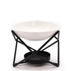 A contemporary oil burner with a glossy white ceramic bowl resting on a black geometric metal frame. The frame has a crisscrossed structure and a lower round holder for a tealight candle. The image is shot on a white background, emphasizing the contrast between the sleek white bowl and the matte black frame.