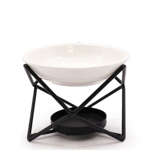 A contemporary oil burner with a glossy white ceramic bowl resting on a black geometric metal frame. The frame has a crisscrossed structure and a lower round holder for a tealight candle. The image is shot on a white background, emphasizing the contrast between the sleek white bowl and the matte black frame.