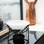 A modern ceramic oil burner featuring a round shallow white bowl resting on a geometric black metal stand with a cross-frame design. The stand has a circular platform underneath designed to hold a tealight candle. The burner is placed indoors on a glossy black surface near a window, with a blurred background showing a wooden decorative vase and natural light.