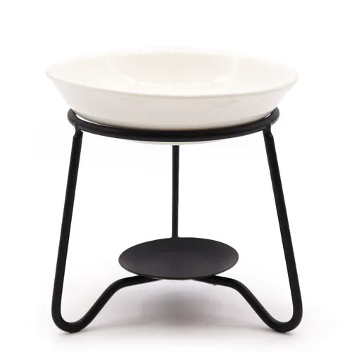A minimalist ceramic oil burner with a white shallow dish supported by a black tripod-style metal frame. Beneath the dish is a flat circular platform for holding a tealight candle. The photo is taken against a plain white background, highlighting the clean and simple design of the burner.