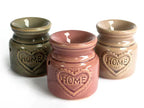 Group of three ceramic oil burners in green, rose, and beige colors, each with a glossy finish and heart-shaped embossed “HOME” design on the front. They are displayed side by side on a white surface.