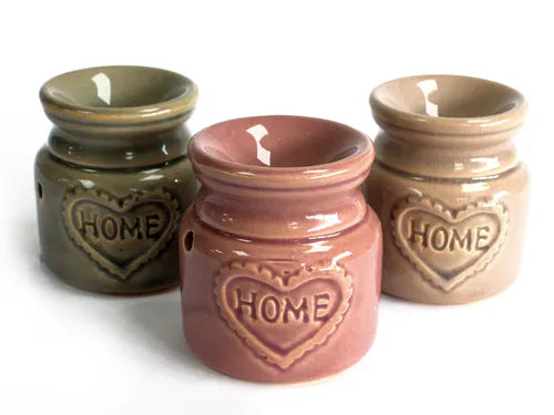 Group of three ceramic oil burners in green, rose, and beige colors, each with a glossy finish and heart-shaped embossed “HOME” design on the front. They are displayed side by side on a white surface.