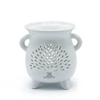 White ceramic oil burner with glossy finish, shaped like a rounded pot standing on three short legs. The front side features a cut-out pattern of a stylized tree of life with spreading branches and a solid trunk, surrounded by small perforations for decorative light effects. Two small rounded handles are placed on either side, and the top has a shallow bowl designed to hold fragrance oil or wax melts.