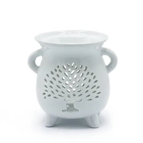 White ceramic oil burner with glossy finish, shaped like a rounded pot standing on three short legs. The front side features a cut-out pattern of a stylized tree of life with spreading branches and a solid trunk, surrounded by small perforations for decorative light effects. Two small rounded handles are placed on either side, and the top has a shallow bowl designed to hold fragrance oil or wax melts.
