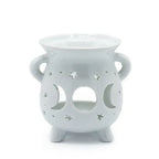 White ceramic oil burner with a rounded cauldron-like shape, two side handles, and three short legs. The front surface is decorated with cut-out shapes of crescent moons and stars, allowing light to shine through when a candle is placed inside. The top has a shallow bowl for holding essential oils or wax melts.