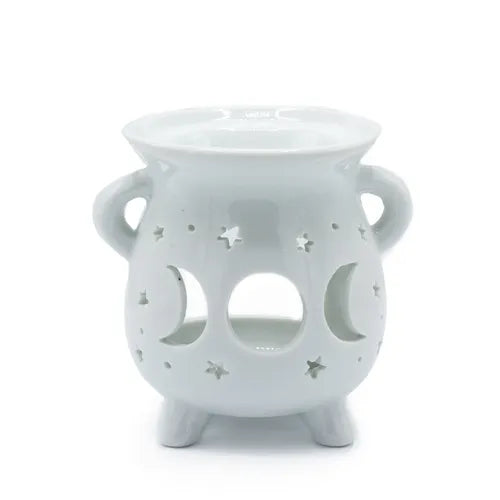 White ceramic oil burner with a rounded cauldron-like shape, two side handles, and three short legs. The front surface is decorated with cut-out shapes of crescent moons and stars, allowing light to shine through when a candle is placed inside. The top has a shallow bowl for holding essential oils or wax melts.