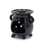 Black ceramic oil burner shaped like a small cauldron, standing on three legs with two side handles. The front has a large cut-out opening with three star-shaped perforations in the back wall, allowing light from a tealight candle to shine through. The glossy surface reflects light, giving it a polished look.