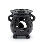 Glossy black ceramic oil burner designed like a cauldron with two side handles and three legs. The front features a large crescent moon cut-out surrounded by multiple star-shaped perforations, creating a celestial theme. The polished surface enhances its decorative appeal while allowing candlelight to project through the cut-outs.