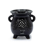 Black ceramic oil burner shaped like a small cauldron with two side handles and three legs. The front surface features a cut-out design of a stylized tree of life with a trunk and many leaf-shaped openings, allowing light to shine through. The top has a shallow bowl for holding essential oils or wax melts.
