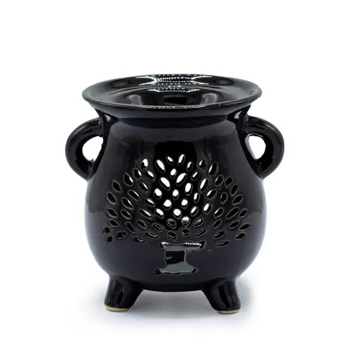 Black ceramic oil burner shaped like a small cauldron with two side handles and three legs. The front surface features a cut-out design of a stylized tree of life with a trunk and many leaf-shaped openings, allowing light to shine through. The top has a shallow bowl for holding essential oils or wax melts.