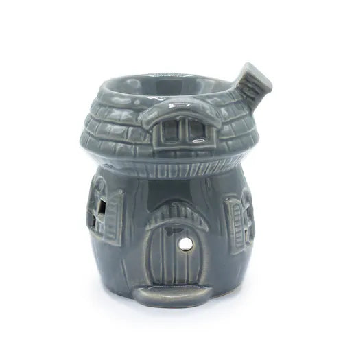 Ceramic oil burner in the shape of a small mashroom-house with a rounded tiled roof and a chimney on the right side. The front has a rounded arched door with a circular cut-out above it, a small dormer window under the roof, and detailed side windows. The top is open to hold fragrance oil. The glossy finish is gray with engraved brick and wood details.