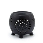 Black ceramic oil burner with a round pot-like shape, raised on four small legs. The surface features intricate cut-out flower of life patterns, including a large central circular flower design with petal-shaped perforations and smaller flower motifs scattered around. The open bowl at the top is intended for holding fragrance oil or wax melts.