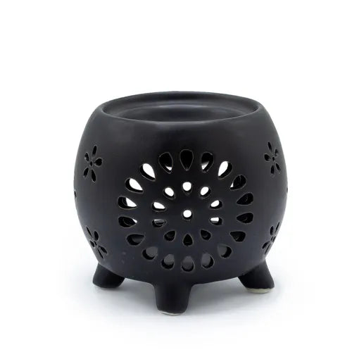 Black ceramic oil burner with a round pot-like shape, raised on four small legs. The surface features intricate cut-out flower of life patterns, including a large central circular flower design with petal-shaped perforations and smaller flower motifs scattered around. The open bowl at the top is intended for holding fragrance oil or wax melts.