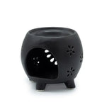Rear view of a black ceramic oil burner, shaped like a round pot with four legs. The back has a large open cut-out to place a tealight candle inside, allowing light to shine through the floral perforations on the surface. The top bowl is slightly recessed, designed for melting wax or holding fragrance oil.