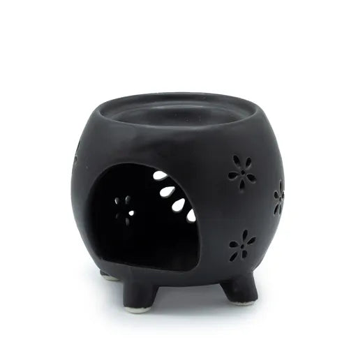 Rear view of a black ceramic oil burner, shaped like a round pot with four legs. The back has a large open cut-out to place a tealight candle inside, allowing light to shine through the floral perforations on the surface. The top bowl is slightly recessed, designed for melting wax or holding fragrance oil.