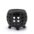 Black ceramic oil burner with a rounded body standing on four short legs, featuring multiple cut-out heart shapes in different sizes arranged symmetrically across the surface, allowing light to shine through. The top has a shallow circular dish for holding essential oils or wax melts.