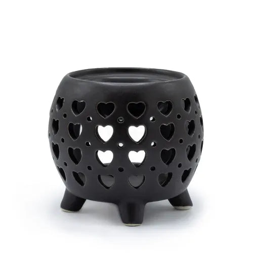 Black ceramic oil burner with a rounded body standing on four short legs, featuring multiple cut-out heart shapes in different sizes arranged symmetrically across the surface, allowing light to shine through. The top has a shallow circular dish for holding essential oils or wax melts.