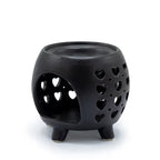 Side view of a black ceramic oil burner with heart-shaped cut-out patterns on the body, placed on four sturdy legs. A large arched opening provides space for inserting a tealight candle inside, while the top has a recessed circular dish designed to hold oils or wax melts.