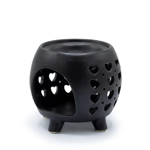 Side view of a black ceramic oil burner with heart-shaped cut-out patterns on the body, placed on four sturdy legs. A large arched opening provides space for inserting a tealight candle inside, while the top has a recessed circular dish designed to hold oils or wax melts.