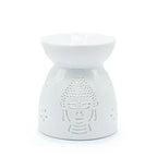 White ceramic oil burner with a wide shallow bowl on top and a perforated design of a Buddha face on the front, surrounded by small floral cutouts.