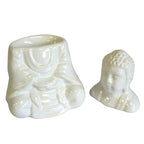 White ceramic oil burner in two separate parts: the hollow base shaped like a seated Buddha body with draped robes, and the detachable Buddha head placed beside it. The glossy surface emphasizes the sculpted folds and facial details.
