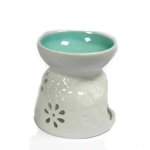 A stunning white ceramic essential oil and wax melt burner with a glossy finish. The top bowl has a beautiful contrasting teal glaze on the inside, providing a pop of color. The burner's body is decorated with elegant, embossed floral patterns and small, flower-shaped cutouts that allow light to shine through. The open side provides easy access for placing a tea light candle. This is an ideal product for home fragrance, aromatherapy, and adding a decorative touch to any room. 