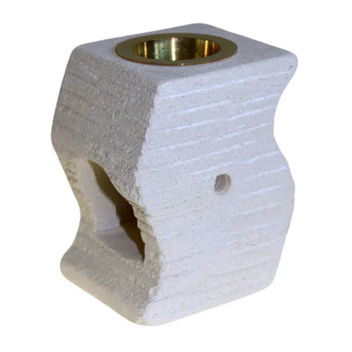 A modern, light-colored soapstone oil burner with a natural, porous texture and a curved, abstract shape. The top is fitted with a polished gold-colored metal bowl for essential oils. The base has a large arched opening on one side for a tea light candle and a small ventilation hole on the other. The soapstone has horizontal grooves carved into its surface, adding to its contemporary, rustic appeal. This essential oil warmer is photographed against a clean white background.