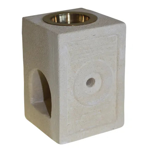 A simple and rustic square oil burner made from light-colored, porous soapstone. The top is fitted with a polished brass bowl for essential oils or wax melts. The base features a large, arched cutout for a tea light candle and a small, circular ventilation hole on the front. A subtle, carved circular design with a line pattern frames the ventilation hole. The product is photographed against a clean white background, emphasizing its natural material and handcrafted design.