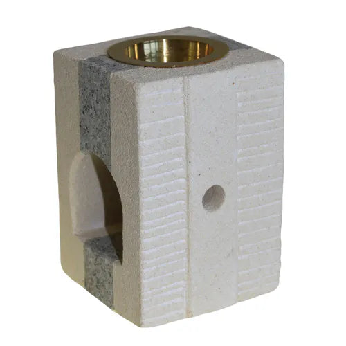 A unique, block-shaped oil burner made from two contrasting types of soapstone. One side is a light, cream-colored stone with horizontal grooves, while the other side is a darker, mottled gray stone. The top of the burner features a polished brass bowl for essential oils or wax melts. The base has a large arched opening for a candle, with the gray stone forming a step at the bottom of the opening. A small ventilation hole is on the side. This oil burner is a great example of rustic, modern home decor.