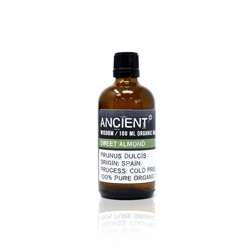 A small, 100ml amber glass bottle of Ancient Wisdom Organic Sweet Almond Oil. The black label is centered on the bottle, featuring the brand name "ANCIENT WISDOM" at the top in white font. Below this is a light green band with "SWEET ALMOND" in white text. The bottom white label provides product details in black text: "PRUNUS DULCIS," "ORIGIN: SPAIN," "PROCESS: COLD PRESS," and "100% PURE ORGANIC." The bottle has a black cap and is photographed against a clean white background.