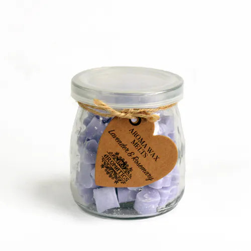A clear glass jar filled with small, light purple, heart-shaped wax melts. The jar is sealed with a lid and tied with a rustic twine bow, from which a heart-shaped brown paper tag hangs. The tag features the text "AROMA WAX MELTS Lavender & Rosemary" in a decorative font, and the product is photographed on a clean white background.