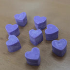 A close-up, overhead view of several light purple, heart-shaped wax melts scattered on a light brown wooden surface. The wax melts are positioned at different angles, showing their detailed texture and slightly rough edges. The wooden surface has a subtle grain visible. The image focuses on the individual wax melts, highlighting their color and shape.