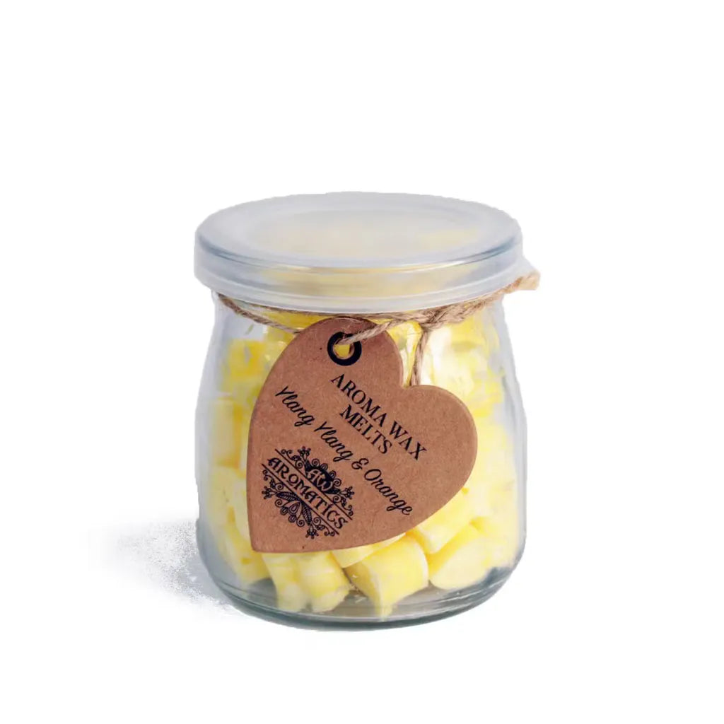 A clear glass jar filled with small, yellow, heart-shaped wax melts. The jar is sealed with a lid and tied with a rustic twine bow, from which a heart-shaped brown paper tag hangs. The tag features the text "AROMA WAX MELTS Ylang Ylang & Orange" in a decorative font, and the product is photographed on a clean white background.