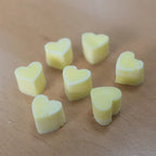 A collection of light yellow, heart-shaped wax melts scattered on a light wood grain surface. The individual melts are shown from a slightly elevated perspective, highlighting their uniform color and solid texture.
