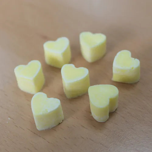 A collection of light yellow, heart-shaped wax melts scattered on a light wood grain surface. The individual melts are shown from a slightly elevated perspective, highlighting their uniform color and solid texture.