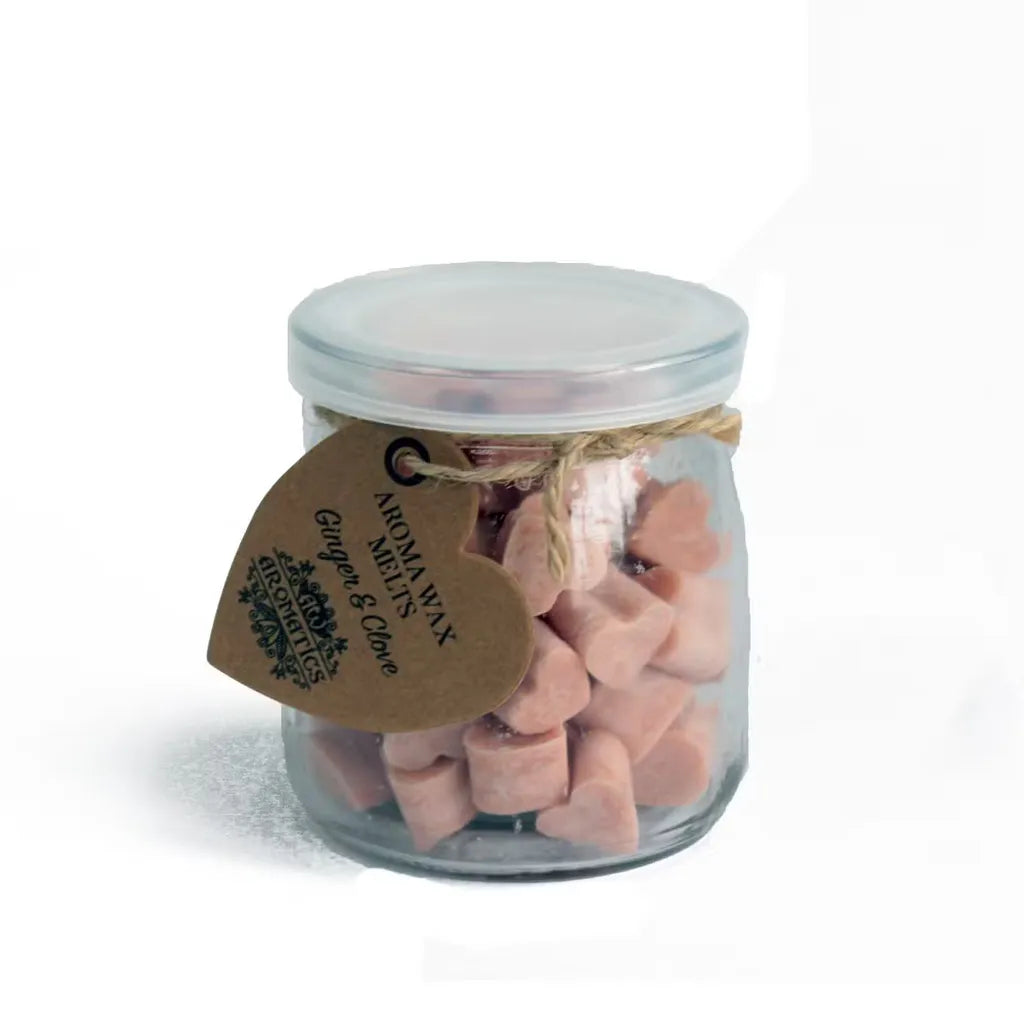 A clear glass jar filled with small, pink, heart-shaped wax melts. The jar is sealed with a lid and tied with a rustic twine bow, from which a heart-shaped brown paper tag hangs. The tag features the text "AROMA WAX MELTS Ginger & Clove" in a decorative font, and the product is photographed on a clean white background.