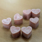 A close-up view of a group of light pink, heart-shaped wax melts scattered on a light brown wood grain surface. The melts have a smooth, solid appearance and are arranged to show their shape and color detail.