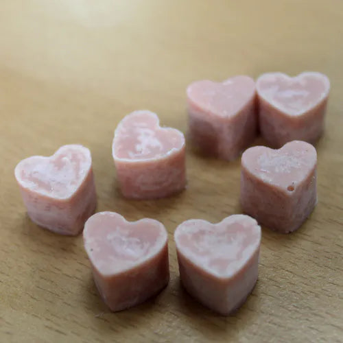 A close-up view of a group of light pink, heart-shaped wax melts scattered on a light brown wood grain surface. The melts have a smooth, solid appearance and are arranged to show their shape and color detail.