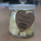 A clear glass jar filled with small, yellow, heart-shaped wax melts. The jar is sealed with a lid and tied with a rustic twine bow, from which a heart-shaped brown paper tag hangs. The tag features the text "AROMA WAX MELTS Nutmeg & Lemon" in a decorative font, and the product is photographed on a light brown wood grain surface.