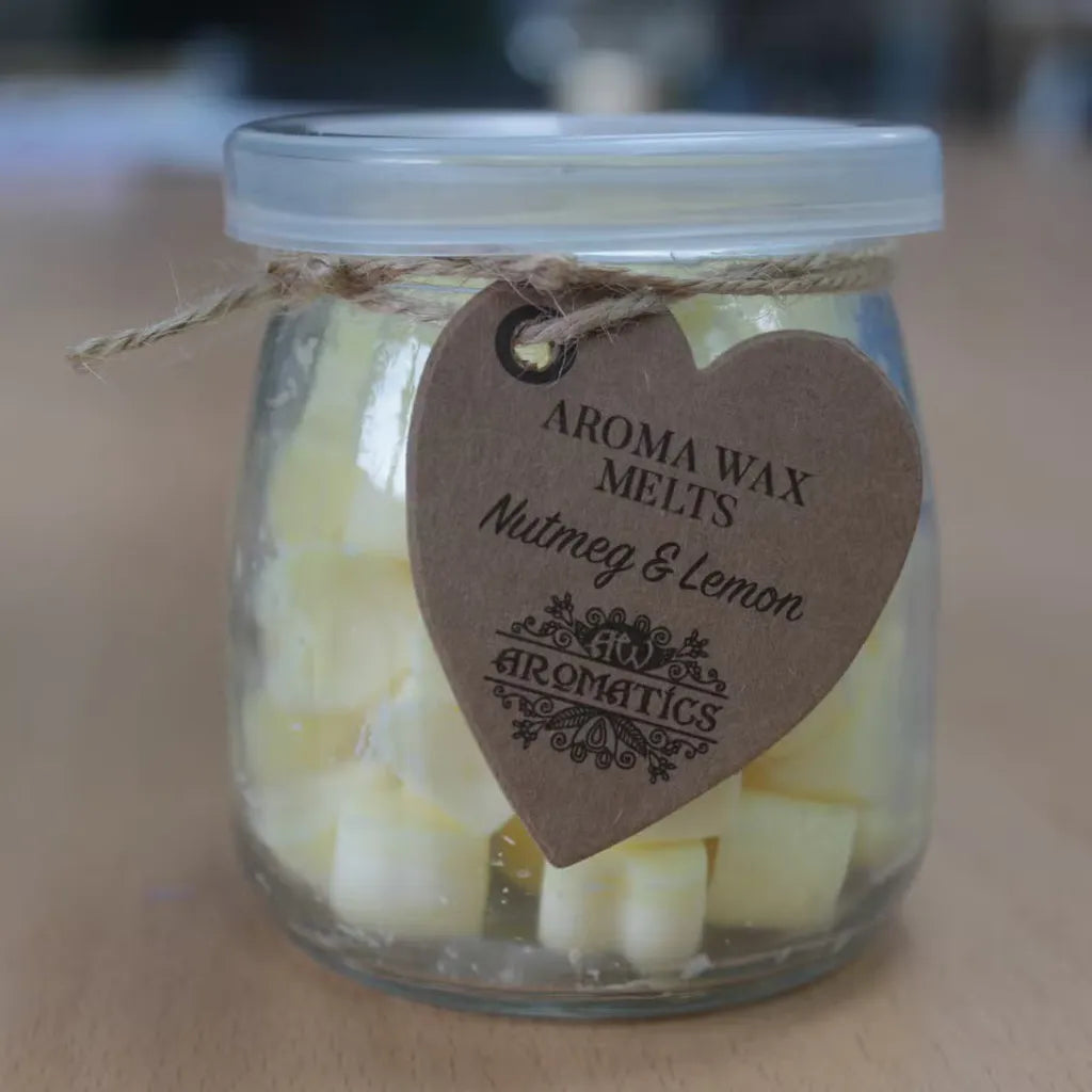 A clear glass jar filled with small, yellow, heart-shaped wax melts. The jar is sealed with a lid and tied with a rustic twine bow, from which a heart-shaped brown paper tag hangs. The tag features the text "AROMA WAX MELTS Nutmeg & Lemon" in a decorative font, and the product is photographed on a light brown wood grain surface.