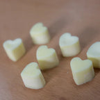 A collection of pale yellow, heart-shaped wax melts scattered on a light brown wood grain surface. The image is a close-up, top-down view highlighting the solid shape and matte texture of the individual melts.