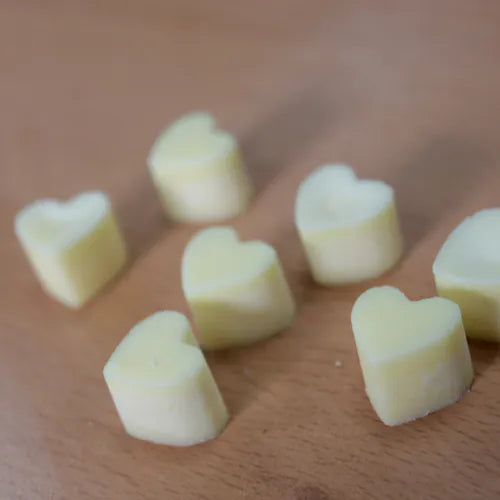A collection of pale yellow, heart-shaped wax melts scattered on a light brown wood grain surface. The image is a close-up, top-down view highlighting the solid shape and matte texture of the individual melts.